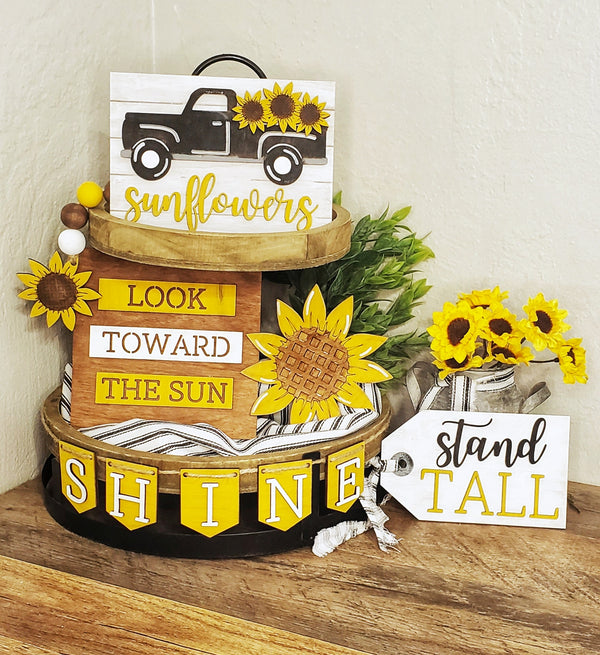 A wooden tiered tray decoration kit with sunflower themes, including cutouts for a truck, sunflowers, and text phrases, arranged on a wooden surface.
